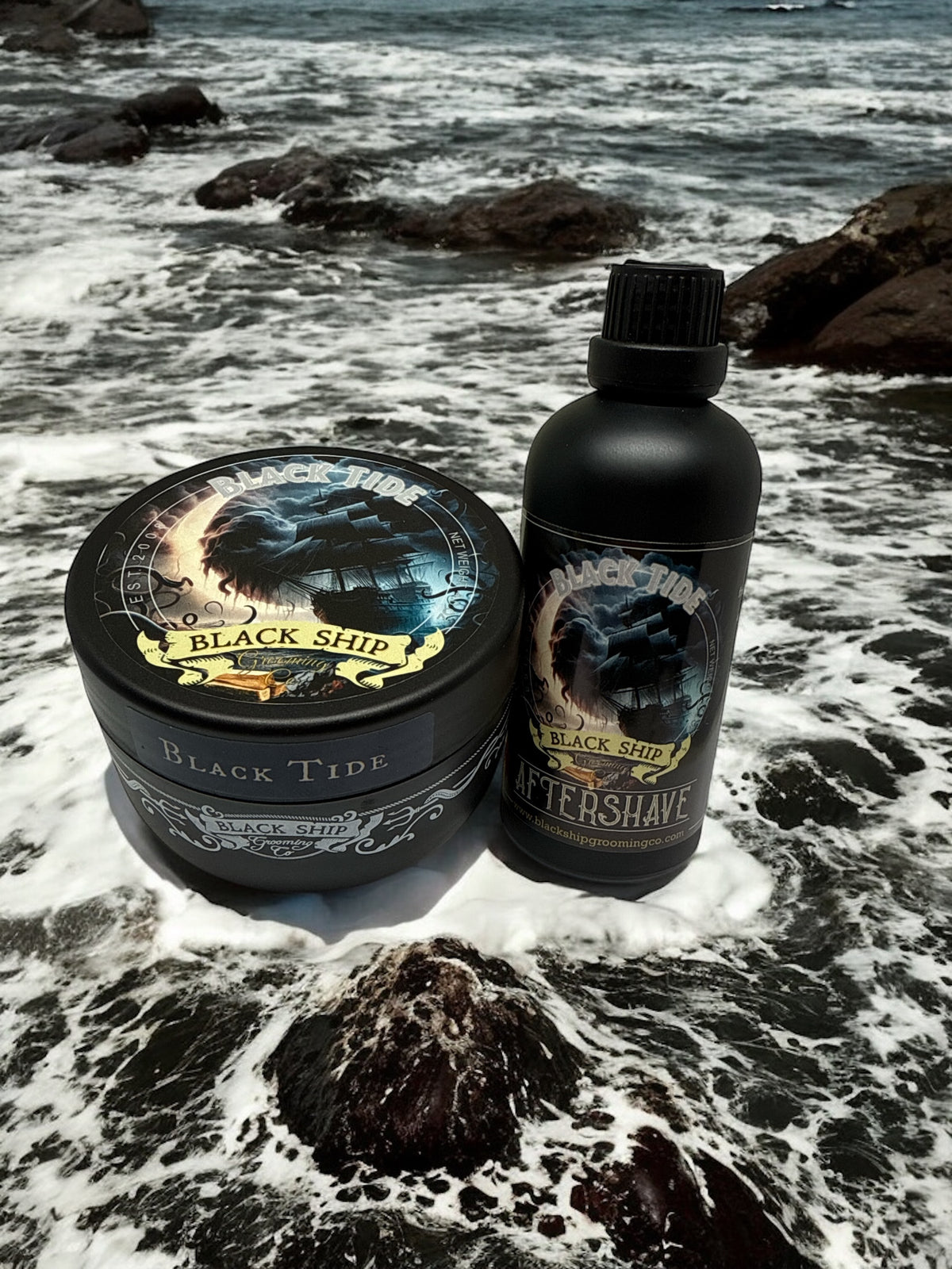bottle of 'Black Tide' aftershave and shaving soap by 'Black Ship grooming co.' on a rocky beach with ocean waves.